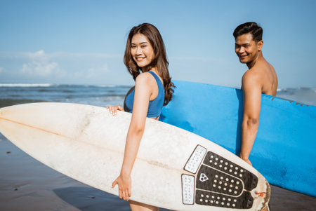 beautiful female surfer and man smiling while looking over with surfboard standing on the beachの写真素材