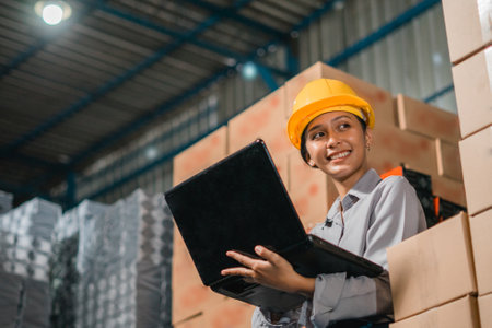 beautiful woman factory worker wearing safety helmet while working with laptop in factory warehouseの写真素材