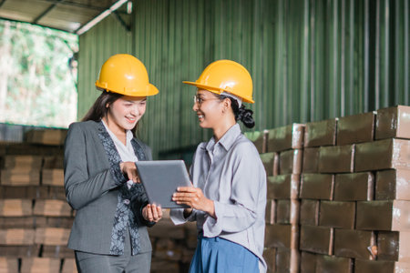 two Asian female factory employees chat while viewing data on a shared tablet in a warehouseの写真素材