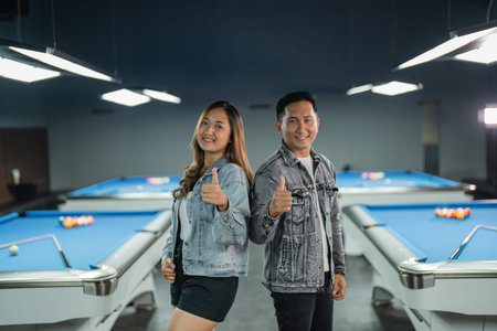 couple of asian billiard players standing back to back with thumb up with the billiard tables at the...