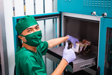male hospital employee wearing uniform and mask with thumbs up while storing blood bag in storage...