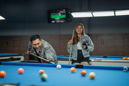 female pool player holding the glass of drink while standing behind the male pool player that poking the white ballの写真素材
