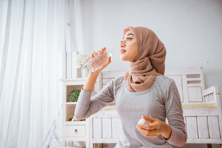 muslim hijab woman smile and drinking from bottle after exercise yoga home workout on orange matress in a roomの写真素材