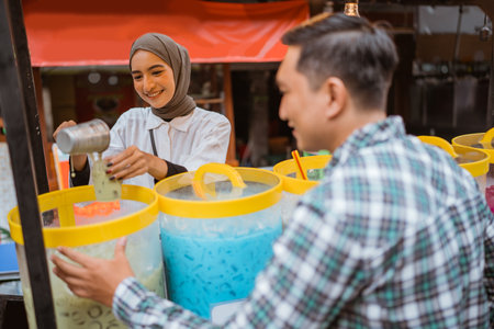 a pretty girl in a veil sells es campur using a scoop to get coconut milk from a jar on a cartの写真素材
