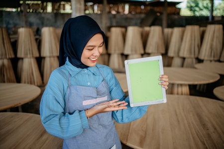 asian female entrepreneur in veil showing tablet screen to camera in wood craft shopの写真素材