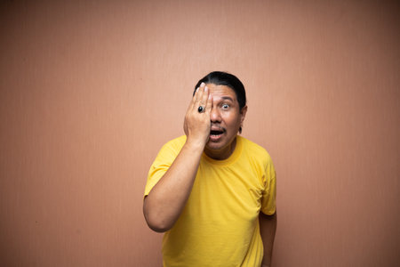 old asian man wearing yellow tshirt covering his half eyes with his right hand in plain background isolatedの写真素材