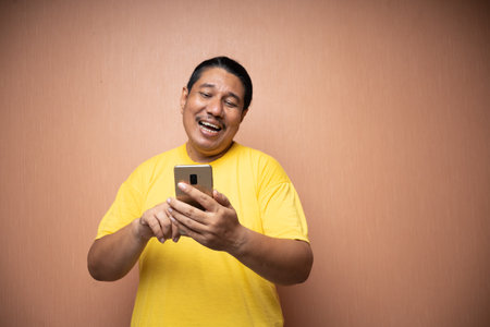 old asian man wearing yellow tshirt holding handphone and using it to texting or video call with each other in plain background isolatedの写真素材