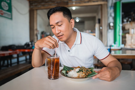 handsome Asian man drinking tea using a straw while having lunch at a dining table in a pecel food stallの写真素材