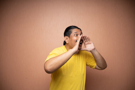 old asian man wearing yellow tshirt with shouting gesture for announcement or sale concept in plain...