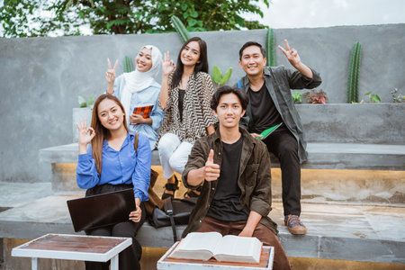 a group of happy students smiling with some hand gestures while sitting together outside the campusの写真素材