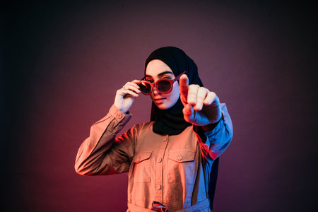 muslim woman wearing black hijab and orange long dress posing sideways and holding sunglasses pointing at cameraの写真素材