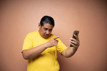old asian man wearing yellow tshirt holding handphone and using it to video call with pointing gesture in plain background isolatedの写真素材