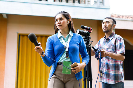 a beautiful reporter in blue cardigan with green name tag hunting for news with a cameraman behind herの写真素材