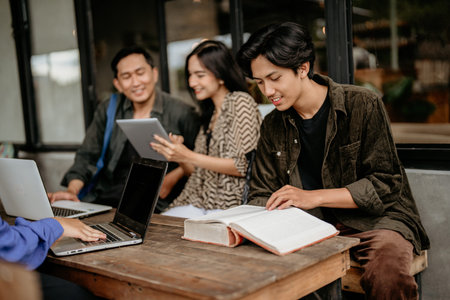 Asian male student reading a thick book while doing coursework at outdoor coworking space