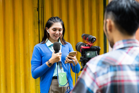 a beautiful reporter in blue cardigan look at her phone while recording videos for news material with the cameraman using one set of camera and a black microphoneの写真素材