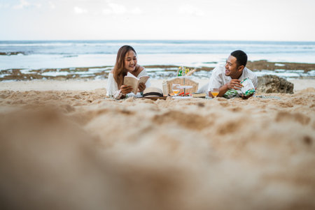 Asian man and woman lying on a picnic blanket while reading a book and joking on the beachの写真素材