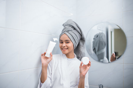 smiling girl in bathrobe holding small bottles of cosmetics in the bathroomの写真素材