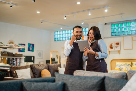 two happy shop clerks as if celebrating something while using a tablet in a furniture storeの写真素材