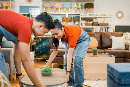 two Asian shop assistants in aprons move a table together while working at a furniture storeの写真素材
