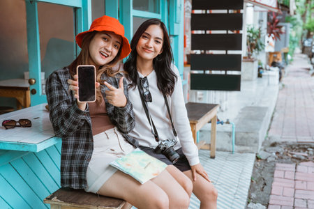 two smiling girls show the blank screen of a cell phone with pointing fingers while sitting at a stallの写真素材