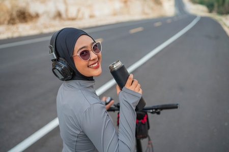 smiling hijab woman holding water bottle when riding bike, biking conceptの写真素材
