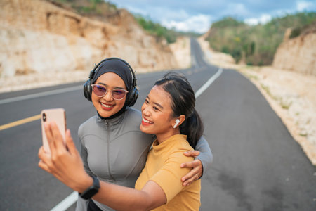 athletic female using phone with hijab friend embracing her, jogging conceptの写真素材