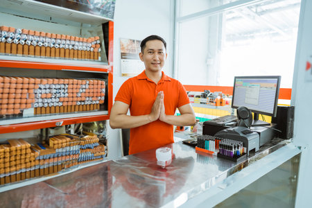 male store clerk smiles at the camera with a greeting gesture at a minimarket cash register