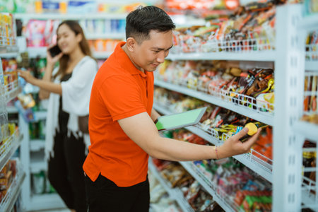 male waiter holding product on shelf while checking with carrying computer tablet in supermarketの写真素材