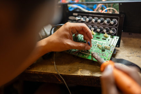 Asian Repairman using soldering iron installs capacitor on printed circuit board at workbench