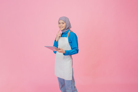 A woman in an apron brainstorms with a tablet, set against a creativity-enhancing pink backdropの写真素材