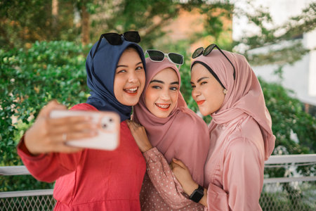 Three joyful women take a selfie in a lush outdoor setting, wearing colorful hijabsの写真素材