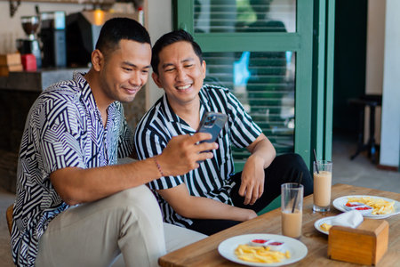 Two close friends sharing a joyful laugh while glancing at a smartphone during a cozy breakfastの写真素材