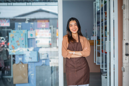 Young asian shopkeeper girl with arms crossed smiling happy standing at the shop