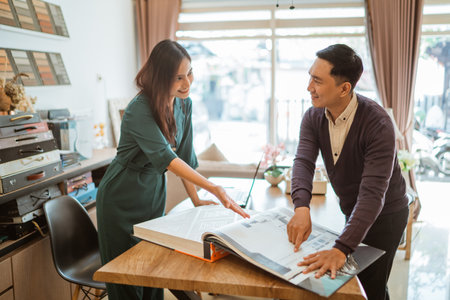 beautiful asian shopkeeper pointing on the catalogue book while explaining the variety of window treatments to the male customerの写真素材