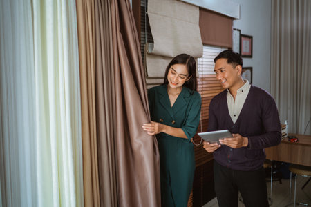 handsome asian customer holding the digital tablet while listening to the female shopkeeper that explaining about the types of curtain fabric at interior shopの写真素材