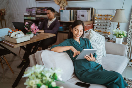 asian beautiful shop owner watching her digital tablet while sitting on sofa with the male shopkeeper at the background inside the interior shopの写真素材