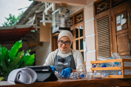 asian woman looking on tablet while cutting plastic bottle for making handmade craft, recycle and reuse conceptの写真素材