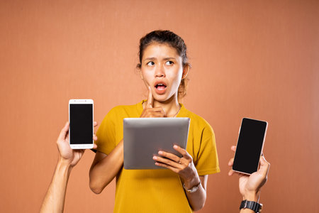 A young woman looks overwhelmed while holding a tablet and two smartphones, showing multitaskingの写真素材