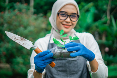 selective focus photo of hijab woman holding green seedling in reused plastic bottleの写真素材