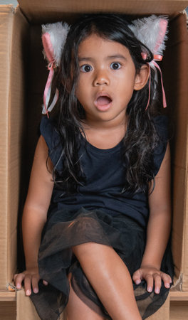 A girl with a surprised expression sits in a cardboard box wearing playful furry earsの写真素材