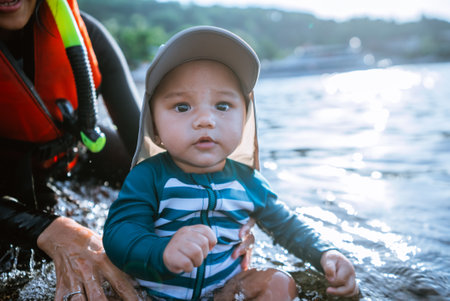 A cute baby in a swimsuit and sun cap plays in the water with a caring adult nearbyの写真素材
