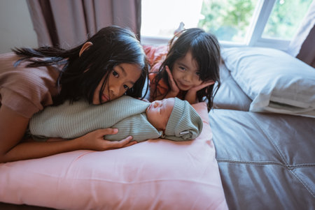 In a cozy bedroom, two loving sisters gaze at their adorable newborn sibling, filled with warmthの写真素材