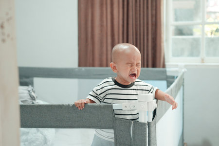 A crying baby stands in a crib, capturing a heartfelt moment in a cozy home settingの写真素材