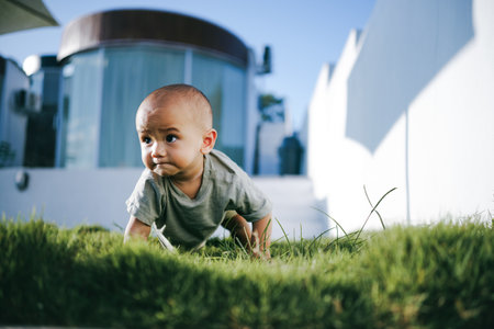 A small baby is happily crawling on the lush green grass in front of a building, enjoying the warmth of the sunny dayの写真素材
