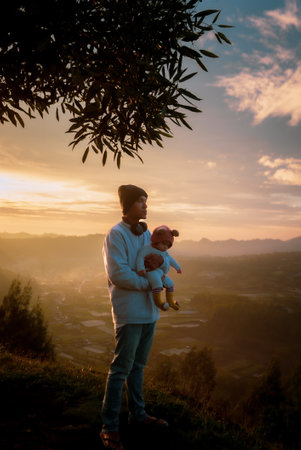 A man is gently holding a tiny baby in his arms, while standing on top of a beautiful hill, enjoying the breathtaking view at sunsetの写真素材