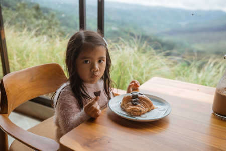 A little girl is peacefully sitting at a small table, happily enjoying a delicious dessert with her fork in hand at that momentの写真素材