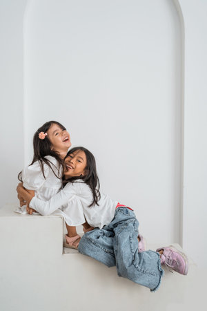 Two little girls, joyfully sitting on the soft floor, are hugging each other tightly, basking in the warmth of their friendship and affectionの写真素材