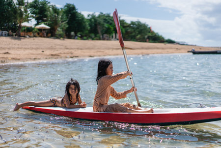 Two girls happily paddling a bright red kayak through the clear ocean waters on a beautiful day, with the sun shining aboveの写真素材