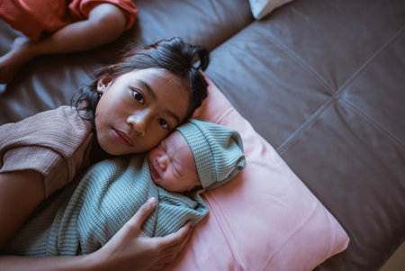 A young girl is gently and lovingly holding her newborn sibling on a warm and cozy sofaの写真素材