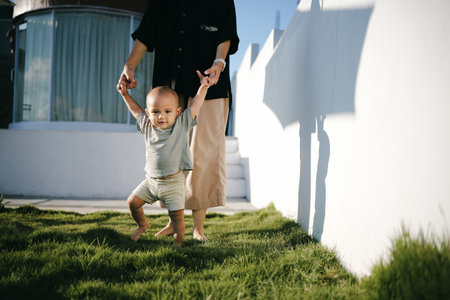 A woman is gently holding a small baby s tiny hand while walking together in the lush green grass on a sunny dayの写真素材
